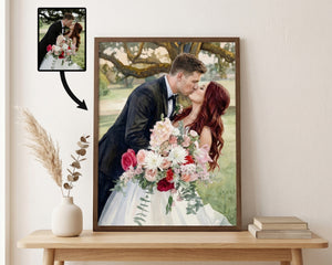 A framed custom wedding portrait print displayed on a light wood console table against a cream wall. The print shows a couple kissing, with the bride in a white gown holding a pink and white floral bouquet.
