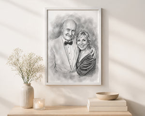 A framed black and white custom portrait sketch of an elderly couple, displayed on a warm beige wall above a light wood console table styled with a vase of baby's breath, a candle, and a small bowl.