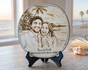A round engraved wooden plaque on a small dark stand, featuring a custom couple portrait with palm trees and beach scenery, plus the text 'Thomas & Rachel ENGAGED MAY 26TH, 2026'. The plaque is photographed on a tabletop near a window with a beach and ocean view in the background.