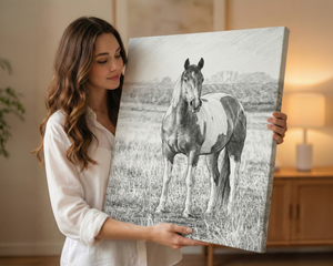 A woman in a white blouse holds up a large square canvas print featuring a black-and-white pencil-style drawing of a horse standing in a field. The scene is set in a warmly lit indoor room with a lamp and wooden furniture in the background.