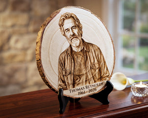 A personalized wood slice plaque featuring a laser-engraved portrait of Thomas Reynolds with the text 'THOMAS REYNOLDS 1961-2026', displayed on a black stand on a wooden table with a blurred home interior background.