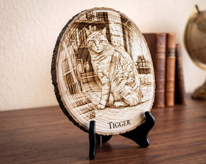 A round laser-engraved wood plaque featuring a detailed portrait of a cat named 'TIGGER' in a library, displayed on a black stand on a wooden table with books in the background.