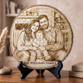 A round laser-engraved wood slice plaque featuring a family portrait and the text 'THE WILLIAMS FAMILY,' displayed on a black stand on a wooden table in a home interior setting.