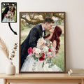 A framed custom wedding portrait print displayed on a light wood console table against a cream wall. The print shows a couple kissing, with the bride in a white gown holding a pink and white floral bouquet.