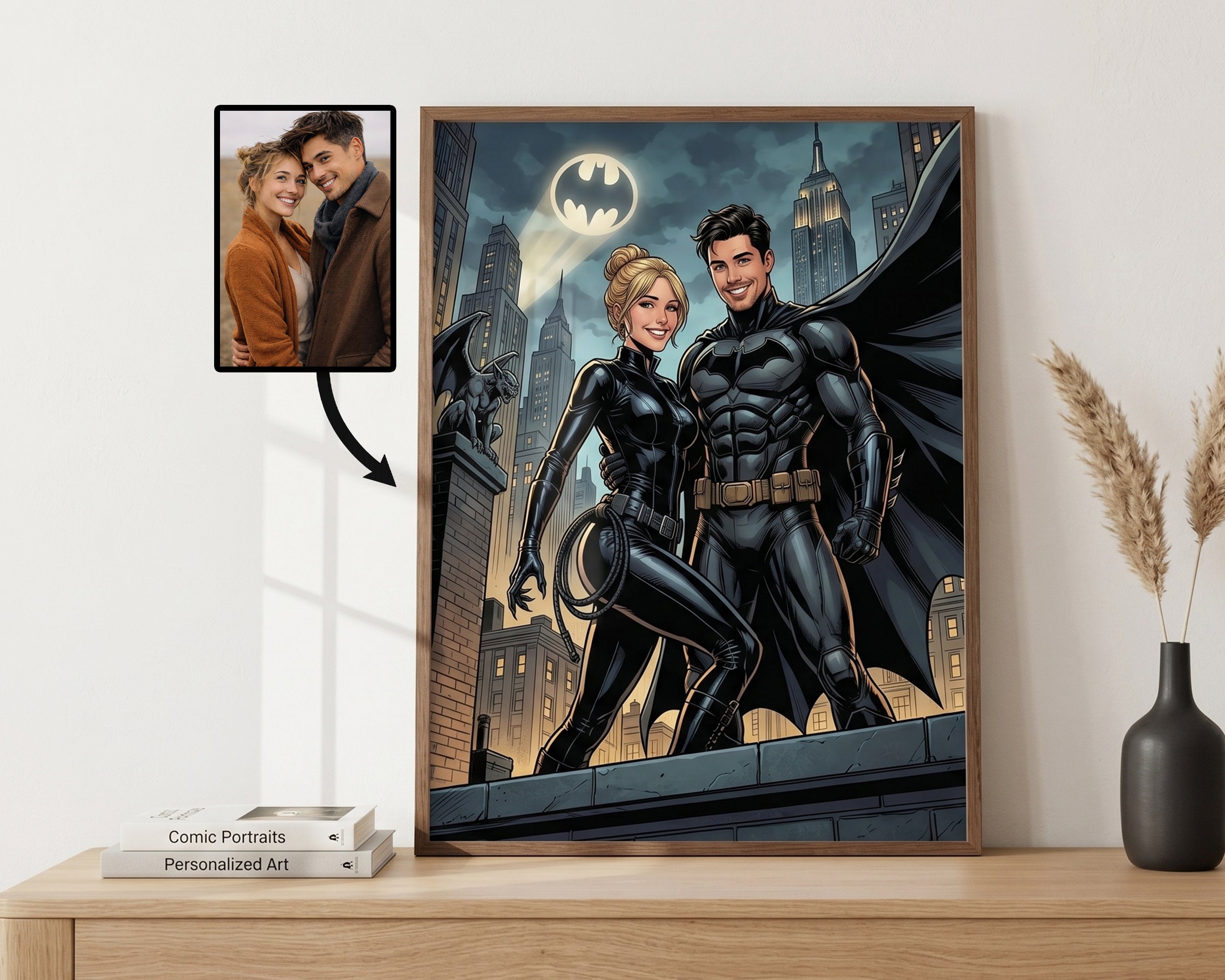 A framed custom comic-style art print of Batman and Catwoman standing on a rooftop at night, displayed on a wooden shelf next to a vase with dried grass and a small inset photo showing the original portrait that was transformed into the artwork.