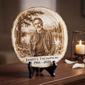 A round engraved wood plaque on a black stand, featuring a portrait of James L. Thompson with the text 'James L Thompson 1961 - 2026' at the bottom, displayed on a wooden table with a lit candle and white flower petals in the background.
