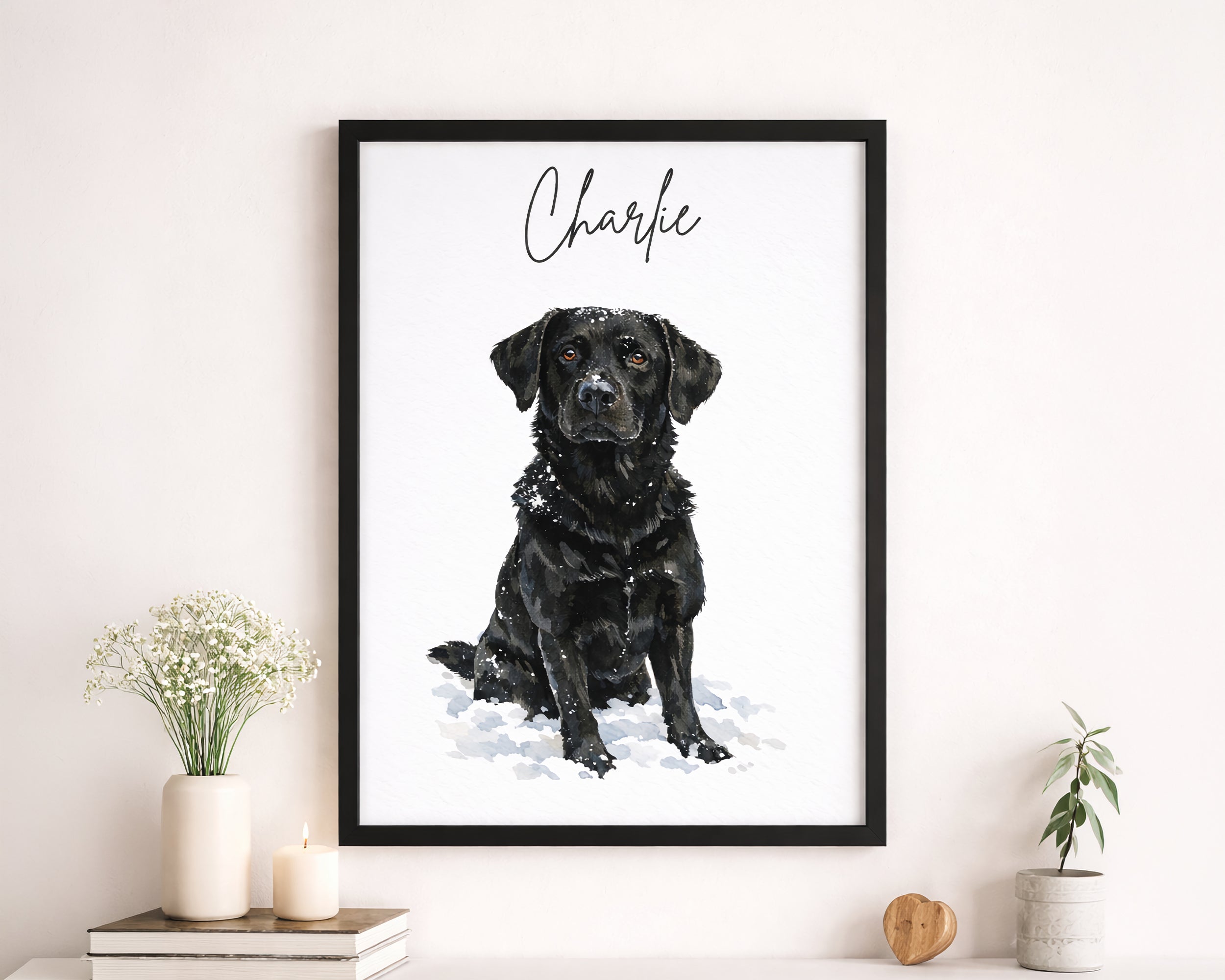 A framed custom pet portrait print featuring a black Labrador retriever named 'Charlie' in a black frame, styled on a white wall above a shelf with books, a candle, and small plants.