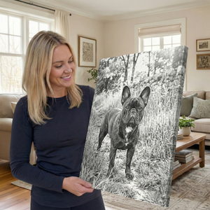 A woman in a navy long-sleeve shirt holds up a large canvas print featuring a black and white photograph of a French bulldog standing in tall grass. The scene is set in a cozy, modern living room with beige walls, framed art, a sofa, and a wooden coffee table.