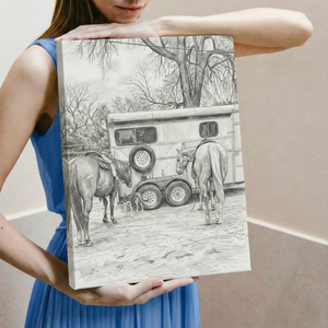 A woman in a blue dress holds up a square canvas print featuring a black and white pencil sketch of two horses and a horse trailer in a wooded setting.