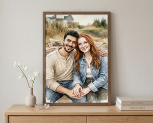 A framed custom portrait painting of a couple sitting together outdoors, displayed on a light wood console table against a neutral wall, with a small vase of white flowers on the left and a stack of books on the right.