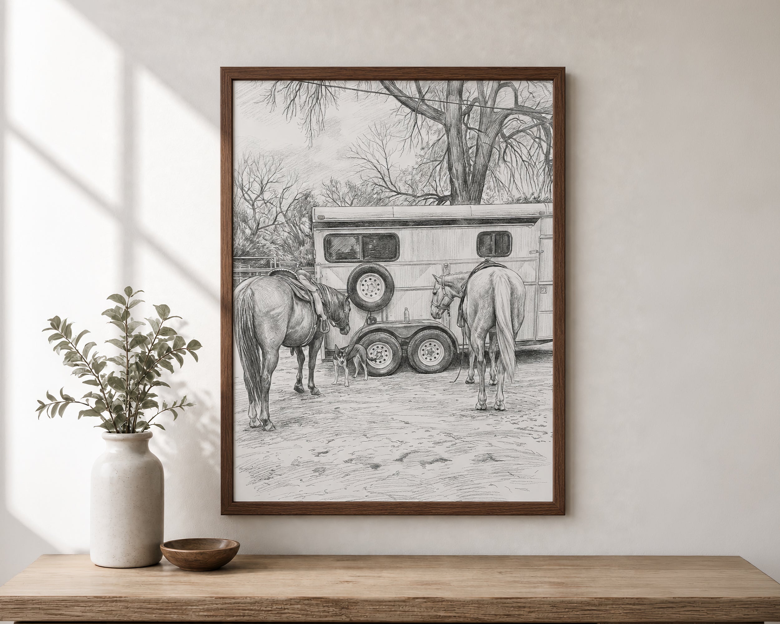 A framed black and white pencil sketch print of horses and a horse trailer, displayed on a wooden console table against a light beige wall with a vase and small bowl nearby.