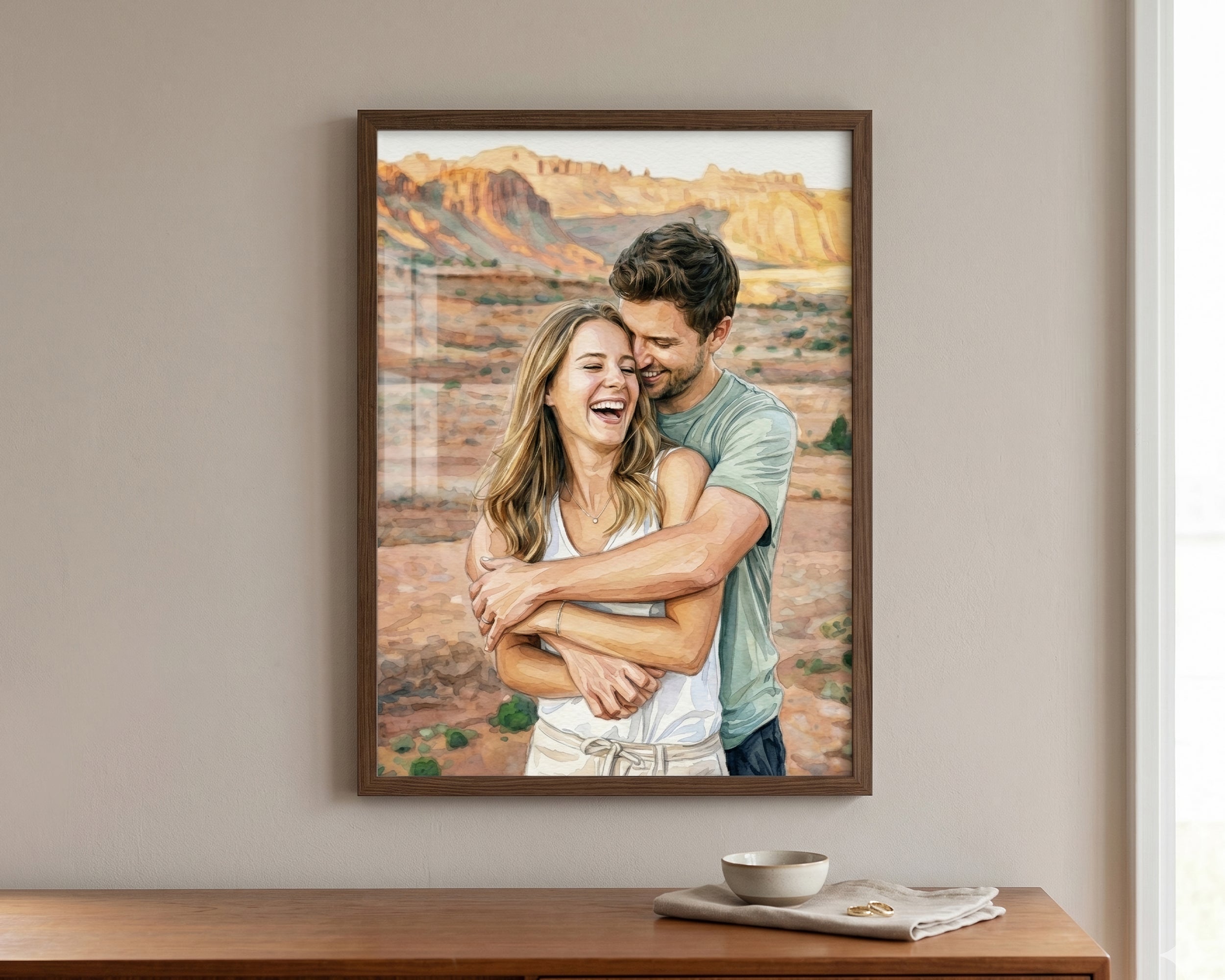 A framed custom photo print of a couple hugging in a desert landscape, displayed on a beige wall above a wooden console table with a bowl and napkin.