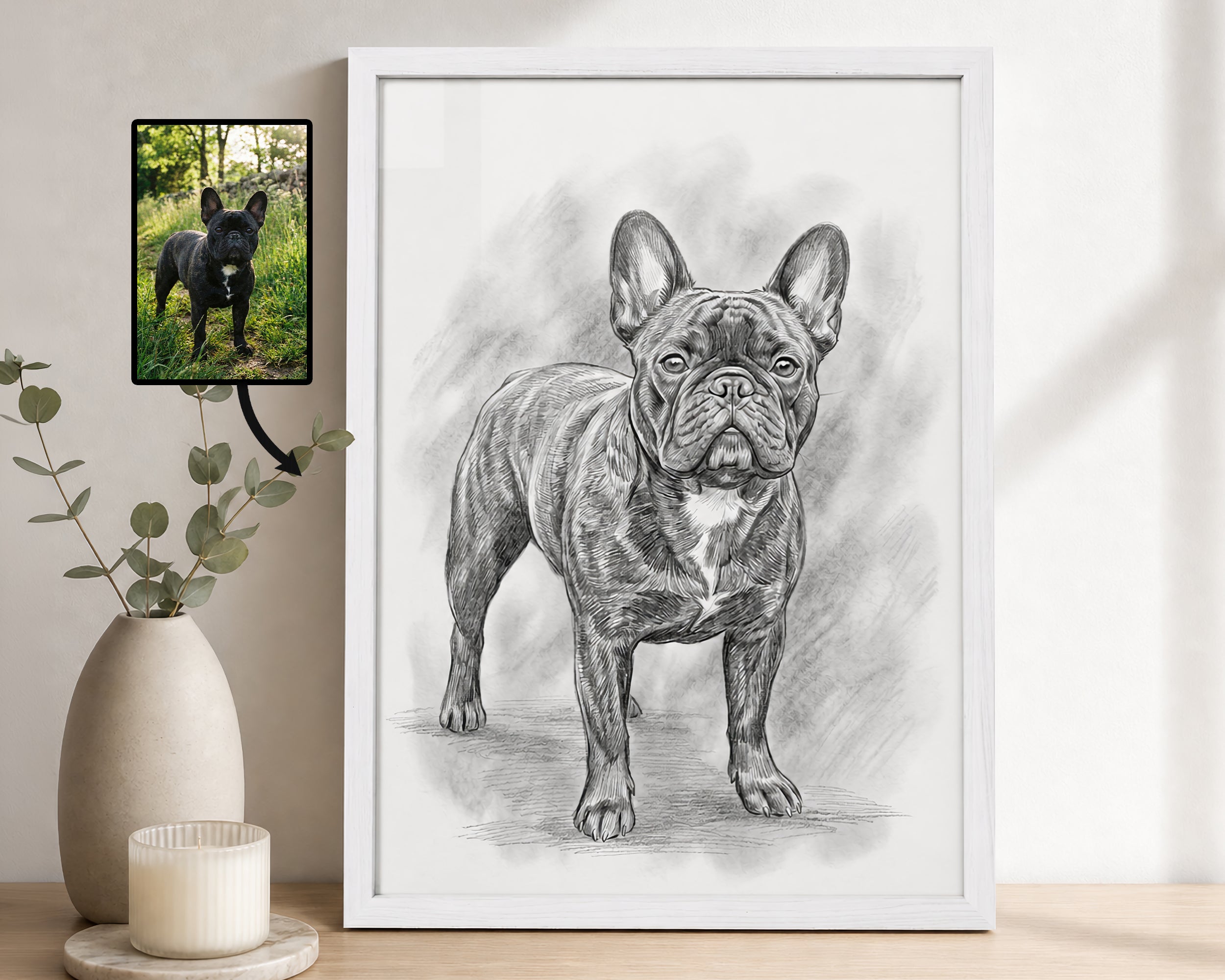 A framed black-and-white sketch portrait of a French bulldog stands on a wooden surface against a light wall. To the left, a small photo of a black French bulldog is shown above a beige vase with greenery and a candle.