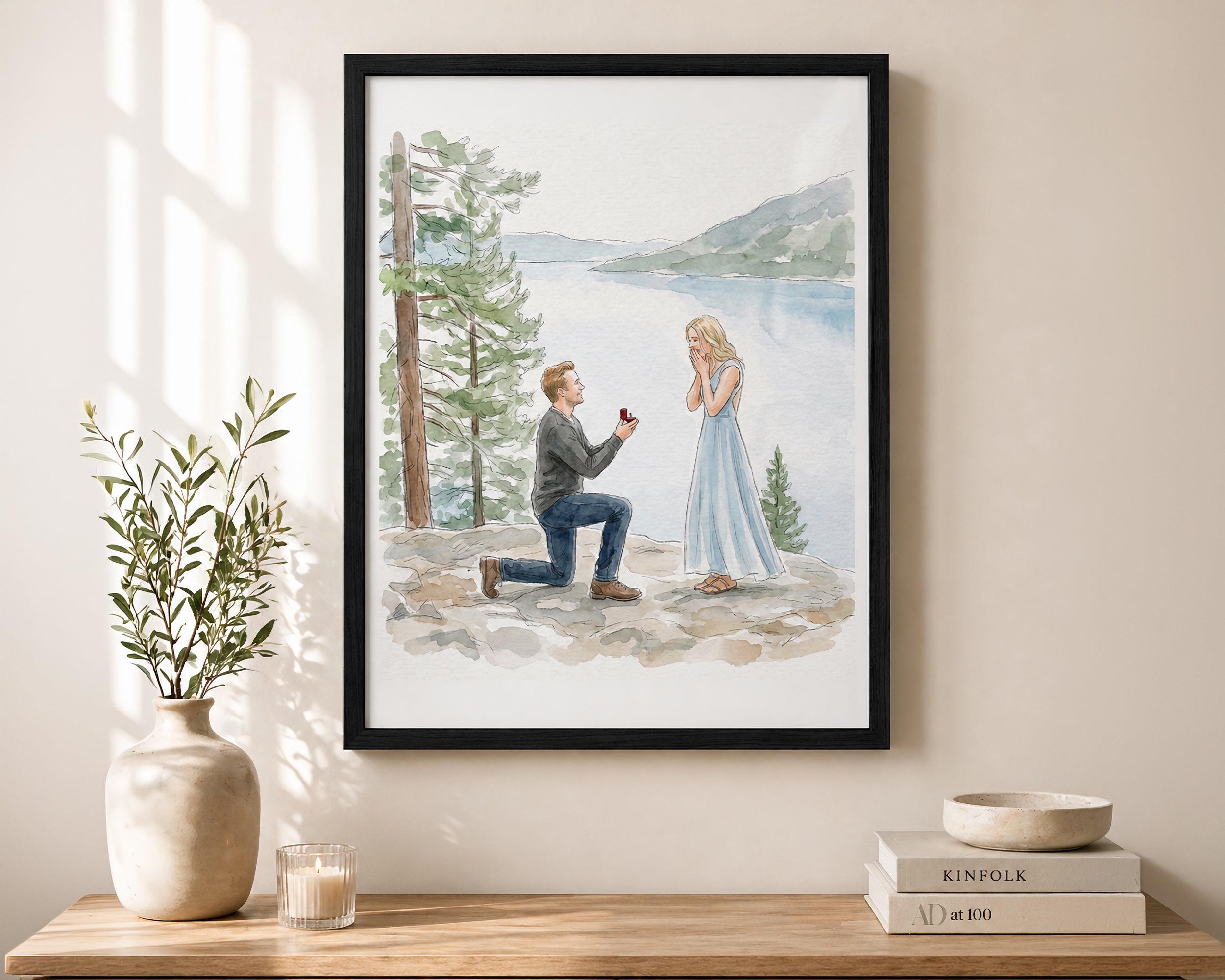 A framed watercolor art print depicting a man proposing on a ring stand to a woman in a blue dress by a lake, displayed on a beige wall above a wooden console table with a vase and books.
