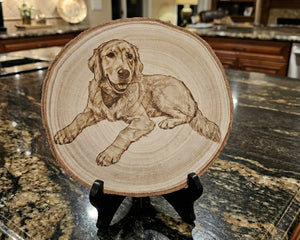 A round wood slice plate featuring a detailed brown engravving or pyrography illustration of a golden retriever dog, displayed on a small black stand on a kitchen countertop.