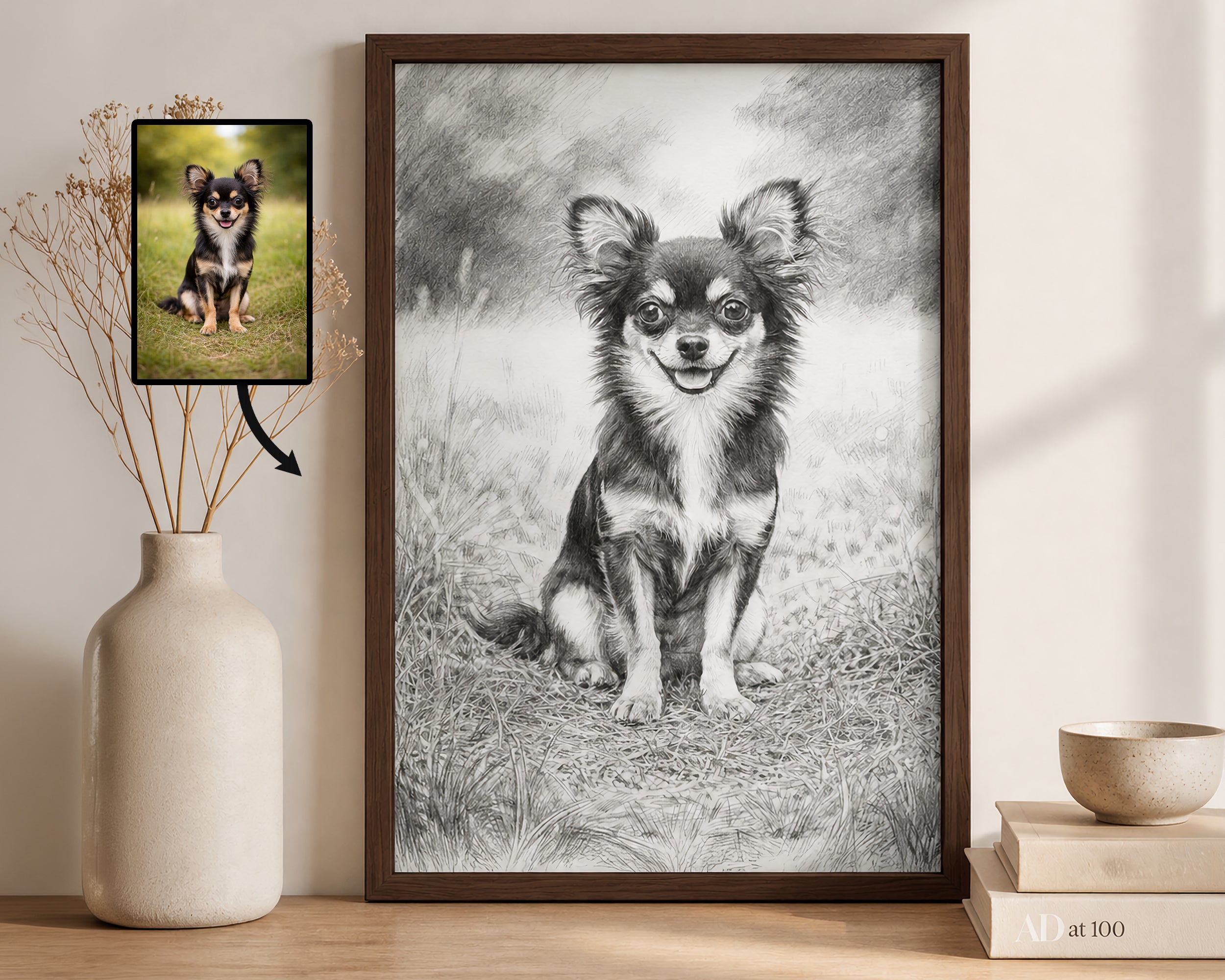 A framed black-and-white portrait print of a chihuahua dog leaning against a wall on a wooden surface, next to a ceramic vase with dried stems and a small inset photo of the dog used as the reference. The print has a dark wood frame.
