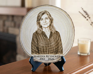 A personalized wooden memorial plaque featuring a portrait engraving of a woman with the text 'MARTHA REYNOLDS 1961 - 2026', displayed on a small black stand on a wooden table in a softly lit indoor setting.