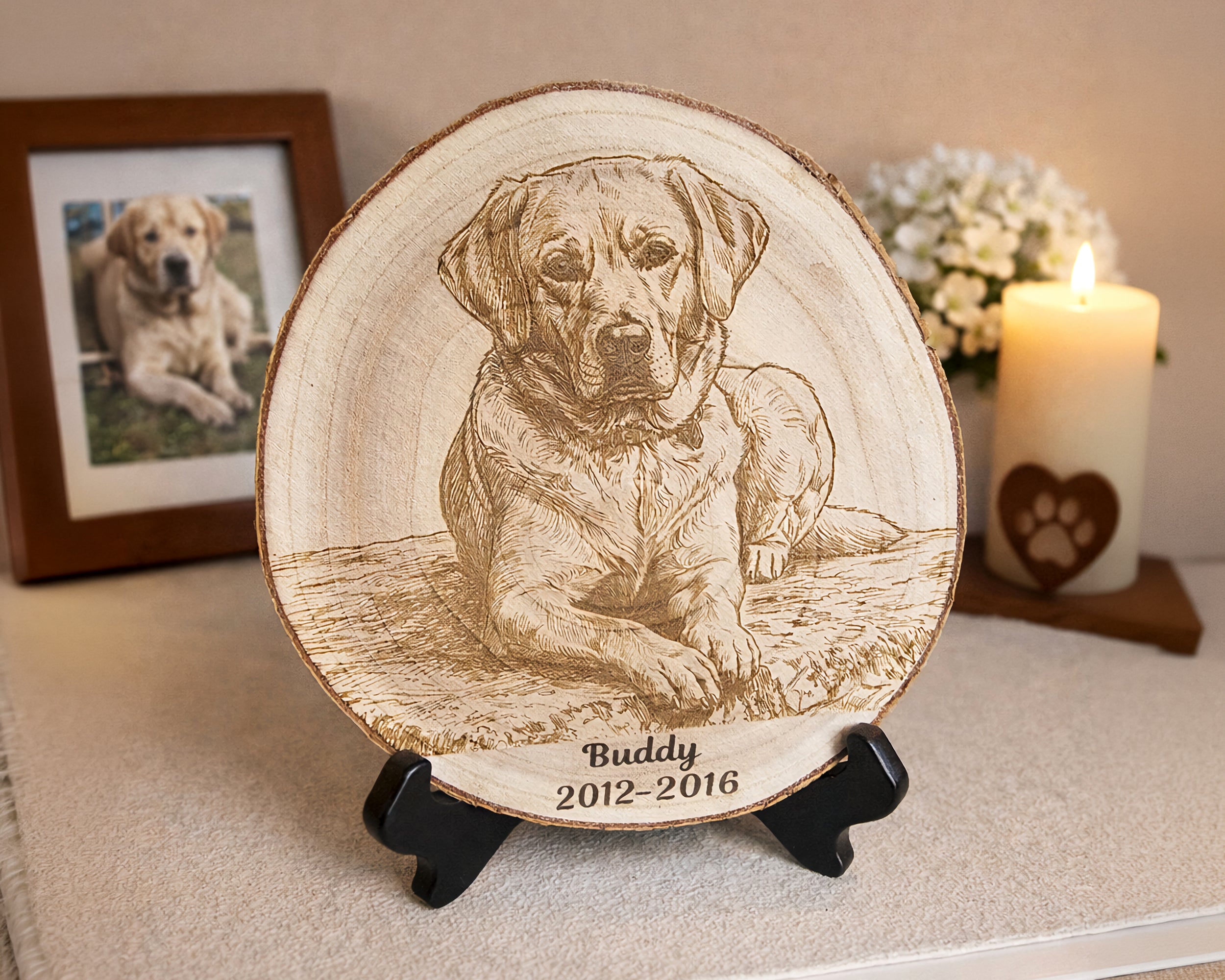 A round wooden plaque featuring a engraved portrait of a dog named Buddy with the years 2012-2016, displayed on a small black stand on a tabletop with a framed photo and a lit candle in the background.