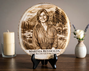 A round wood slice plaque featuring a portrait engraving of Martha Reynolds and the text 'MARtha REYNOLDS GONE BUT NEVER FORGOTTEN,' displayed on a black stand on a wooden surface with a candle and small vase in the background.