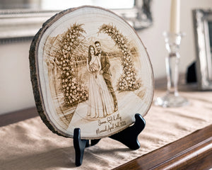 A round natural wood slice plaque with a laser-engraved wedding portrait of a couple under floral arches, displayed on a black stand on a table runner in a home setting.